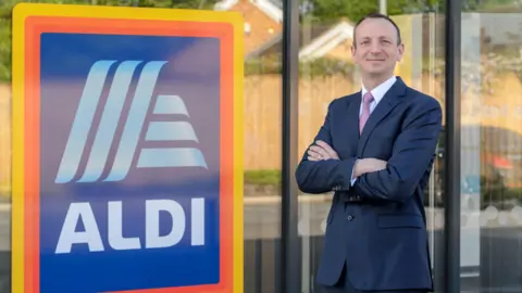 A man in a navy blue suit stands with his arms crossed next to a large sign for Aldi. 