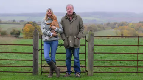 Farm Fest A woman with long blonde/brown hair and wearing a grey jumper is next to a man in a coat. They are both wearing blue jeans and standing by a gate in a field.