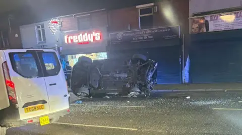 Handout An overturned car lies on Hyde Road - only its underside is visible. Shattered glass can be seen in the road next to it.