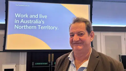 Jessica Bayley/BBC A photo of Geoff Totham stood in front of a projector which reads 'Work and live in Australia's Northern Territory'.