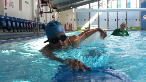 Ruth swims in a pool as her instructor looks on.