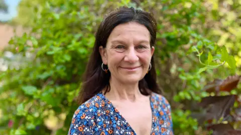 A smiling Bridget Smith looks at the camera. She wears a blue, orange and white flowery top. In the background are some trees with green leaves. 