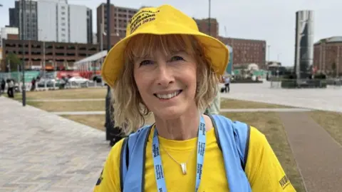 Sharon Hackett, a woman with blonde hair, a yellow hat, a yellow t-shirt and a blue bib. There are various buildings in the background, which is located near Grand Central Station in Belfast.