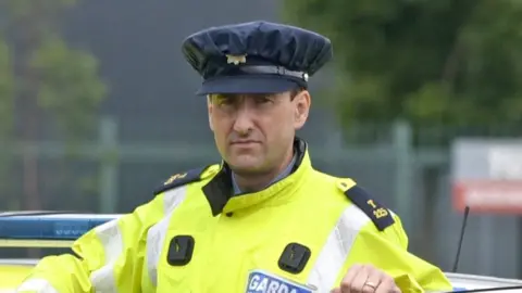 A man wearing an Irish police officer uniform of a yellow hi-vis jacket standing behind the open door of a police car which is stationary.