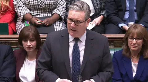 PA Media Sir Keir Starmer, a man with grey hair and glasses wearing a dark grey suit and blue tie, speaks while standing in front of green leather-seated benches occupied by members of Parliament in the House of Commons.