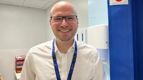 A smiling man with a bald head and glasses in a white shirt with a blue lanyard in a room with white walls and a blue curtain