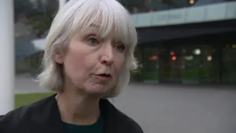 Tracey Lazard, a woman with short grey hair wearing a dark coloured blazer and t-shirt. She is looking at a reporter off-camera and City Hall buildings are visible in the background