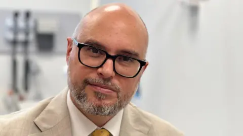 Medical Oncology Consultant Dr Carles Escriu wears a beige suit with a white shirt and yellow tie with blue spots. With close cropped hair, he wears dark-framed glasses.