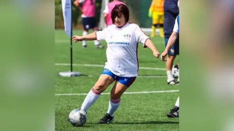 Annie Booth Annie Booth has short red/brown hair. She is wearing a white football top with Sport Parkinson written on it. She had blue shorts and white socks. She is kicking a football. She is on a football pitch. There are other players in a blurred background.