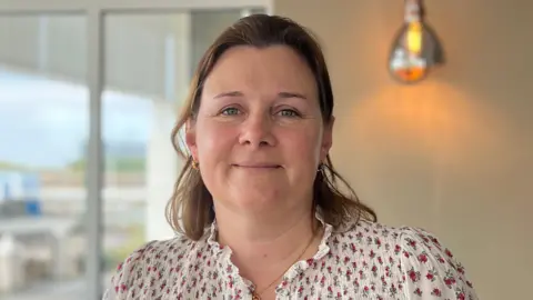 Fiona Kerley facing the camera. She his wearing a white floral top and a necklace, and has medium length brown hair.