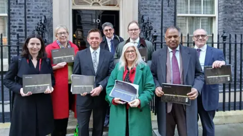 Eight people stood outside number 10 Downing Street, holding copies of a black booklet. The three women and five men are all wearing coats and jackets and smiling at the camera.