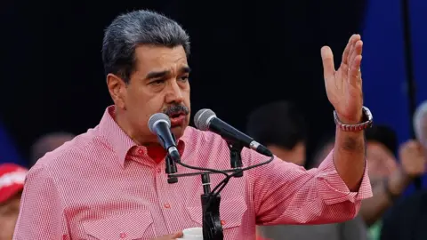 Venezuelan President Nicolás Maduro wears a red shirt and speaks into a microphone. He is gesturing with his hand. 