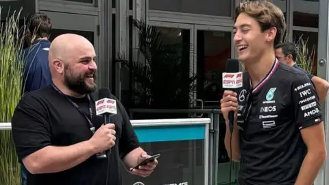 NATE SAUNDERS Nate Saunders (left) laughs while holding up a microphone. Mercedes F1 driver George Russell (right) also laughs while holding a microphone.