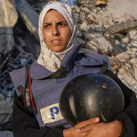 AP A woman wearing a headscarf and a press tabard carried a helmet and camera strap with rubble behind her