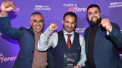 Three men from Whitefield Youth Association, dressed smartly, smiling and raising their fists in triumph