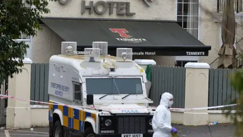 Pacemaker A Police Service of Northern Ireland is parked behind red and white police tape in front of the entrance to the Greenvale Hotel. Behind the land rover is a grey fence, which is in front of an awning under a sign in silver letters saying 'Greenvale Hotel'. In the foreground of the picture, in front of the police land rover, is a forensic officer wearing white protective gear from head to toe and purple gloves