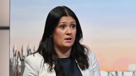 Lisa Nandy, who had long dark hair, appears animated in a picture that seems to be taken mid-conversation. She is wearing a white blazer and is sat in a TV studio. 