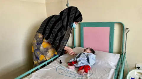 Aakriti Tapar / BBC Zamooda, y pañuelo en la cabeza y su negro y transporte, usan máscara de rampa, con pantalones. Él tiene un tubo en la nariz y pequeño.