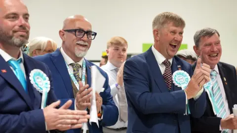 Reform UK activists wearing suits with blue and white rosettes smile and clap as they celebrate results being announced in Essex.
