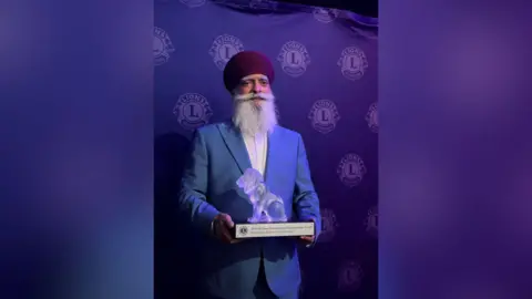A man wearing a red turban and a blue blazer with a white shirt is holding a trophy that's in the shape of a lion that is clear.