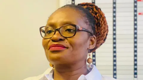 Sibongile Tshabalala Sibongile Tshabalala, wearing a white blouse, lipstick, glasses and dangly earring with her hair in a bun, smiles.