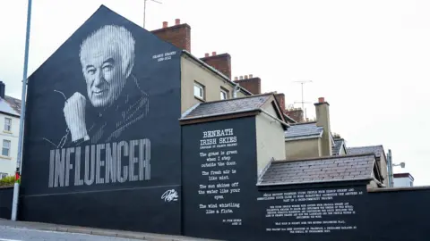 Féile Media A gable wall of a terraced house with a mural painted upon it. It features a portrait of the pet Seamus Heaney pictured above the word influencer. The mural is painted in only black and white. The words of a poem are also written on the wall