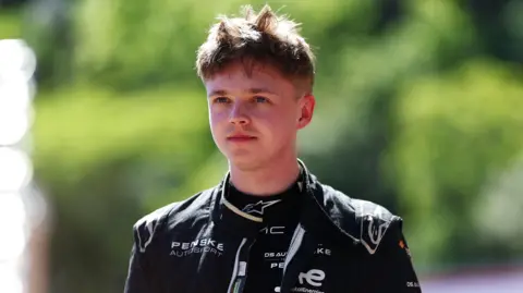 Taylor Barnard wears a black racing suit. He is taking an earpiece out of his ear and looking off into the distance. 