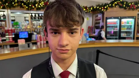A young man who is standing in front of bar at a theatre. He is wearing a white shirt and a black waistcoat as well as a red tie. The bar in the background is decorated with Christmas garland and lights.