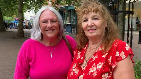LDRS A woman with grey hair and a pink jumper, standing in a high street next to another woman with long ginger hair and a red dress with a white floral pattern. Both are smiling at the camera.