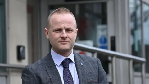 Mr Bryson is looking at the camera. He has no expression and has short light hair. He is wearing a grey suit and purple tie. 