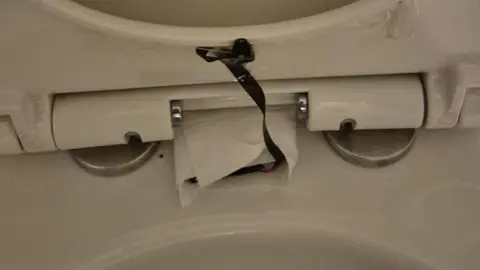 The lifted lid of a white toilet seat, with a black camera tucked under its rim and a wire leading to another part of it wrapped in kitchen roll
