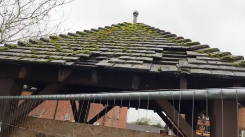 The roof of a pagoda with loose and broken tiles, covered in moss. There is grey metal fencing seen blocking it off.