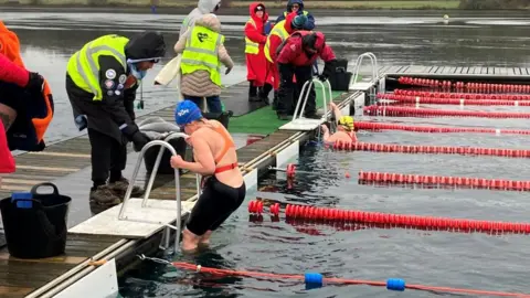 BBC/Mark Ansell A swimmer wearing a blue swim cap with white text, an orange top, and black swim bottoms is climbing out of the water using a metal ladder attached to a dock.
The swimmer is emerging from a lane marked by red floating lane dividers, which are arranged in parallel lines across the water.
A black bucket is placed on the dock near the swimmer.
Another swimmer, wearing a yellow swim cap, is still in the water near a ladder, holding onto the dock edge.
Several individuals are standing on the dock, wearing high-visibility yellow vests and warm clothing.