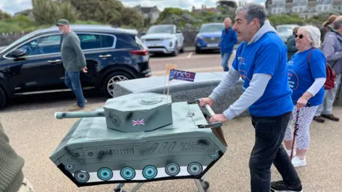 A man pushing a shopping trolley which has a cardboard tank mounted on it.