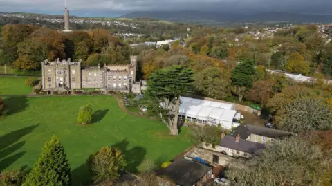 Manxscenes The Nunnery from above. The site comprises of large green grassy areas, an old grey building with turrets. A white marquee has been erected on the grounds. Lots of trees and houses can been seen in the background.