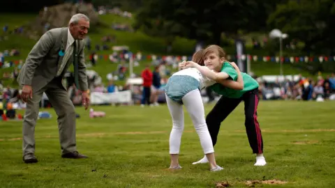 Getty Images Two young girls with blond hair, facing each other and leaning forwards, with their upper bodies in a wrestling hold. Each has their chin on the other's shoulder, with arms around the other's upper back, hands gripped together. One girl is wearing traditional white long johns and white top, with turquoise velvet trunks. The other has a green top and black jogging bottoms, with triple red strips down the sides. They're on a grassy field with out-of-focus spectators and bunting in the distance. Near them is an older man, acting as referee and leaning forward to see clearly, who's wearing a moss green tweed suit and tie.