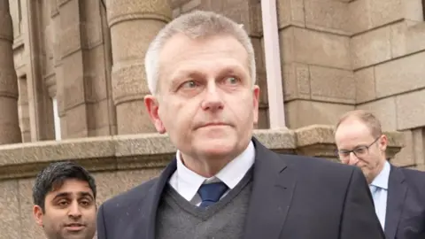 BBC Deputy Philip Ozouf is outside Jersey's Royal Court. He's wearing a suit and is looking away from the camera. He is being followed by two men in suits, one with a microphone.