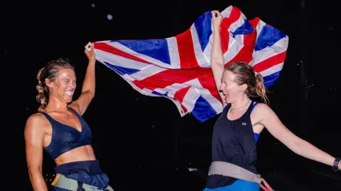 The picture shows two rowers celebrating on the deck of a boat at night. One of them is holding a large Union Jack above her head, letting it billow out behind her, while the other stands beside her with an arm raised in triumph. Both look exhausted but jubilant, as if they have just completed a major challenge at sea. 