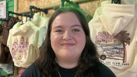 A young woman is looking directly at the camera, and smiling. She has long dark hair and blue eyes, and her bottom lip is pierced. She is wearing a black t-shirt and a long silver necklace. She is standing in a shop in front of cream coloured hoodies hung on the wall with surfing pictures on them.