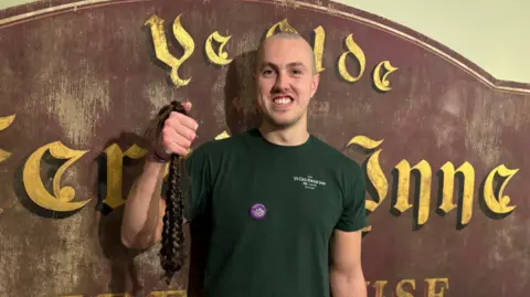 A man with a closely shaved head is holding a large piece of dark plaited hair in one hand. He is smiling