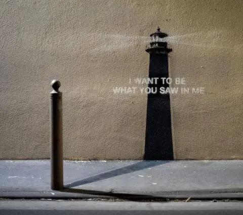 Banksy/Instagram A photo of the Banksy piece of art. A metal bollard is in the foreground on the pavement, while a painting of a lighthouse is on the wall superimposed with a quote "I want to be what you saw in me".