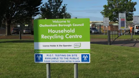 A green sign for Swindon Road Recycling Centre in Cheltenham that reads "Welcome to Cheltenham Borough Council Household Recycling Centre". The sign is on a grass verge at the front of the site. The recycling centre can be seen in the background along with a car park and men in orange hi-vis clothing in a van.