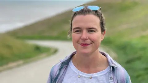 A woman with sun glasses on her head is smiling and standing in front of a grassy slope with the sea in the background.