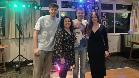 Family A family of four stand with their arms around each other while standing on a dance floor with disco lights behind them.