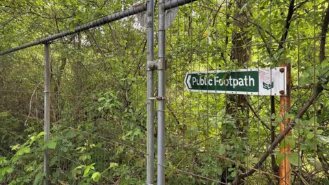 BBC/Lucy Ashton There is a public footpath sign in woodland with a high metal fence in front of it