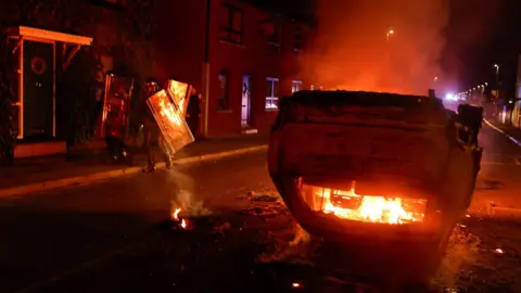 Reuters Police officers near a burning vehicle during a second night of riots in Ballymena
It is night time, a car is on its roof burning.
The officers are in black riot gear and holding plastic shields