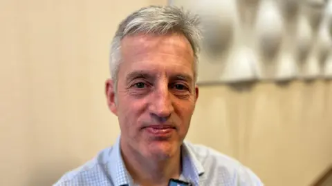 Jim Savege looking directly at the camera with a slight smile. He is wearing a light blue checkered button-down shirt with a small clip-on microphone at the collar