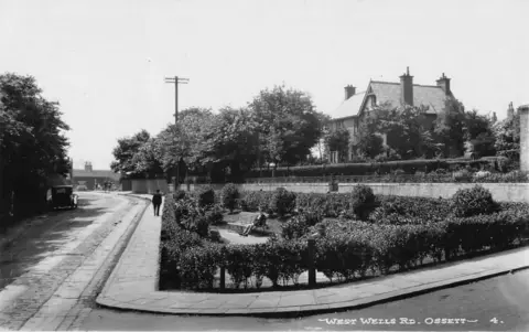 Ossett Through the Ages A black-and-white photograph of a road showing a quiet street scene from the past. In the foreground, there is a paved pavement bordered by cobblestones. There is small garden area enclosed by hedges, with a person sat on a wooden bench. Behind the garden are tall trees and greenery, which partially obscure a large house.