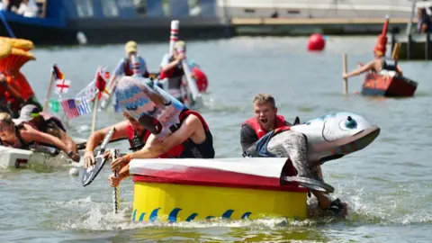 Plaster/Bristol Harbour Festival Four men are in a cardboard boat on the water, you can see more people in cardboard boats in the background.