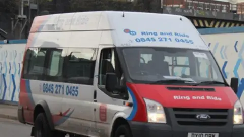 A white Ford mini bus with a red bonnet and blue writing which says "Ring and Ride" and a telephone number. 